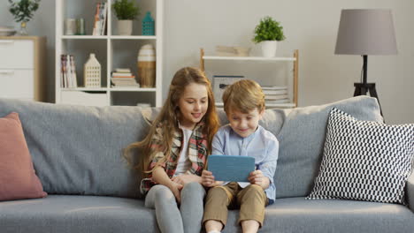 lindos niños pequeños sentados en el sofá y viendo algo en la tableta, luego su madre viene y los abraza