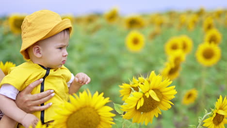 cute frowning little child in yellow panama looking at sunflowers. mom's holding her son looking straight ahead. blurred backdrop.