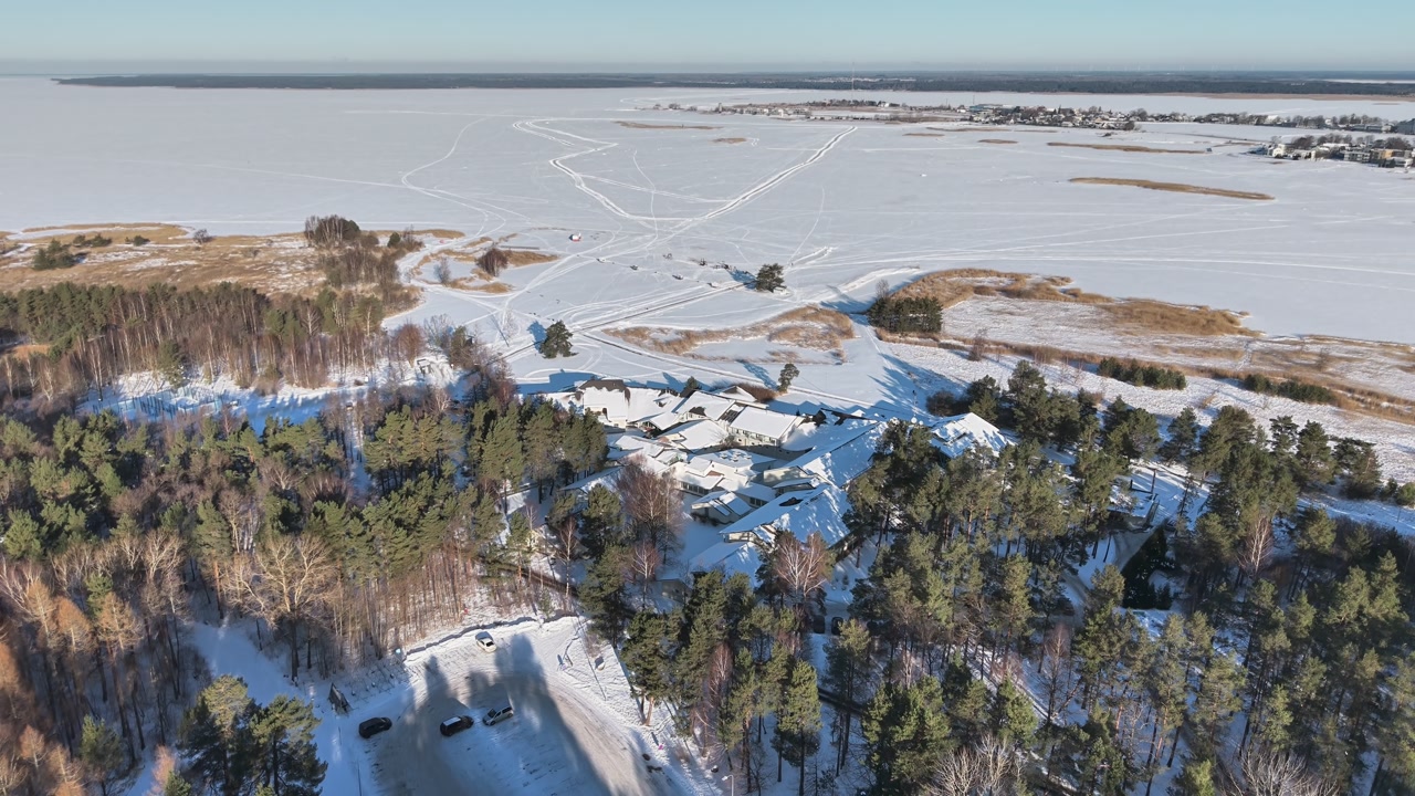 Drone aerial over snowy forest and frozen coastline near Haapsalu with scattered houses and winter roads, a peaceful countryside scene ideal for travel and seasonal backgrounds