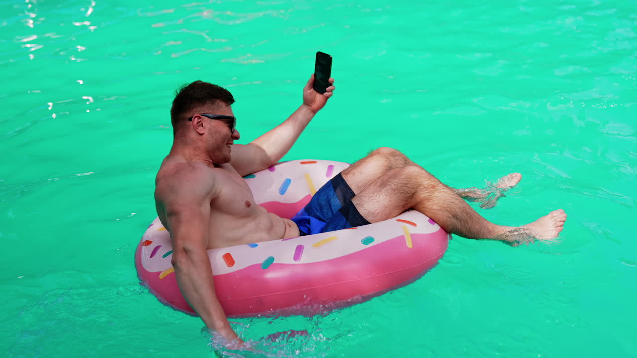 Muscular male sits in the inflatable circle holding a phone. Man taking selfie while resting in the pool.