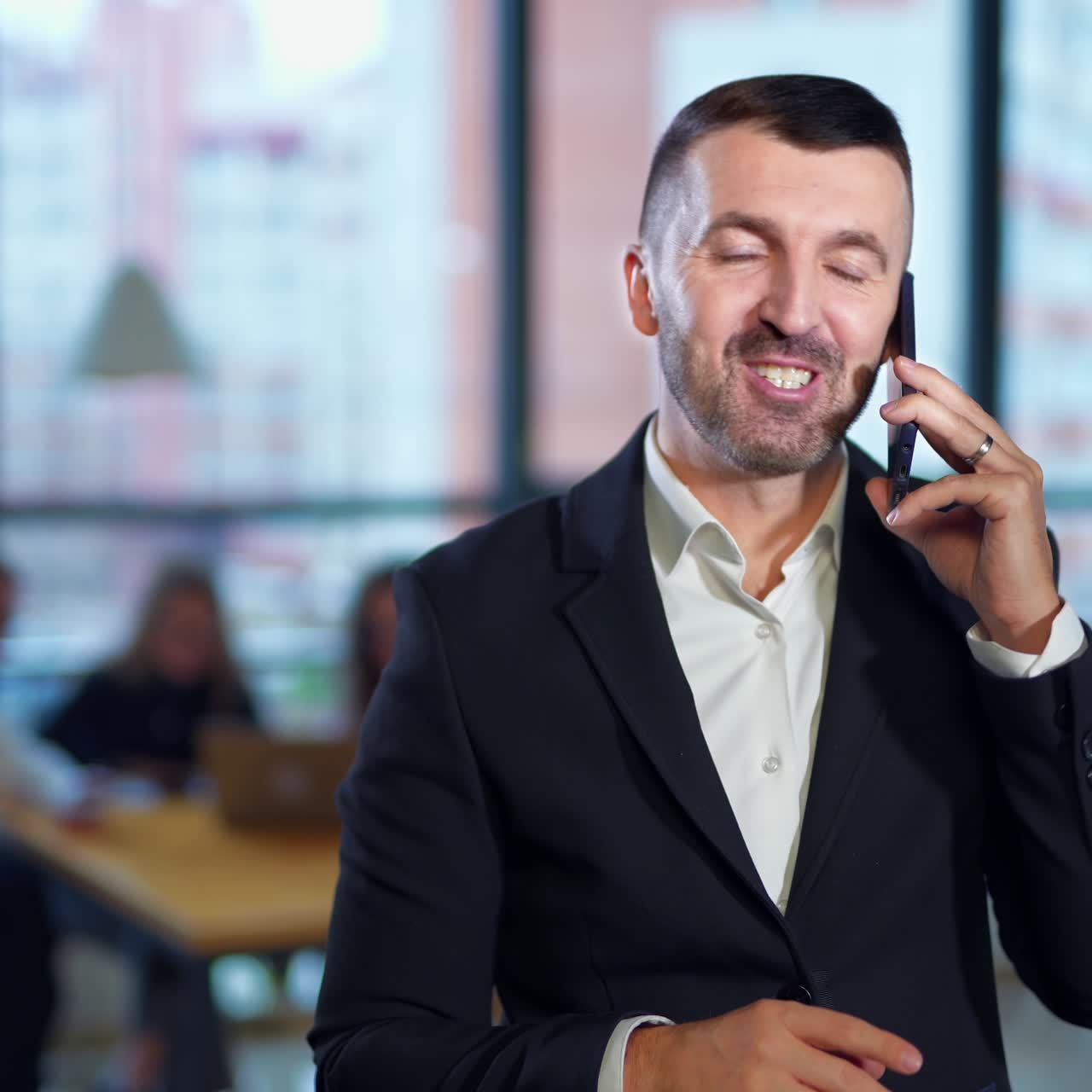 Uomo d'affari barbuto, allegro e positivo che parla al telefono di fronte alla telecamera. Staff dell'ufficio sullo sfondo sfocato