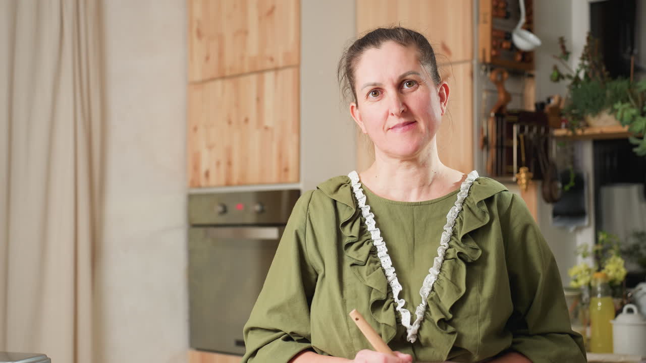 Cook in green gown with lace trim standing in cozy kitchen, holding spatula with head gently tilted, smiling softly, background includes wooden cabinets, electric oven, plants, and kitchen tools