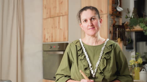 cook in green gown with lace trim standing in cozy kitchen, holding spatula with head gently tilted, smiling softly, background includes wooden cabinets, electric oven, plants, and kitchen tools