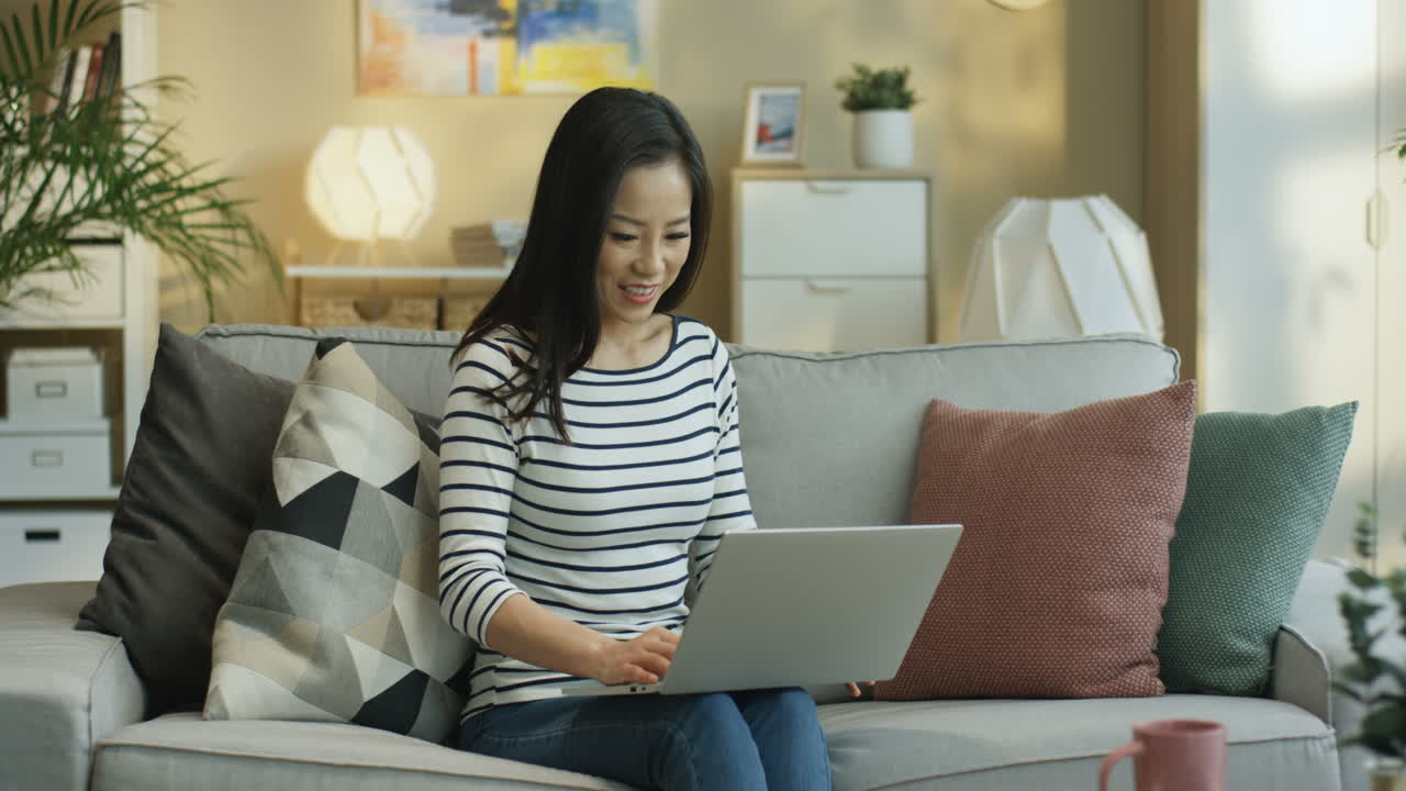 줄무늬 블라우스를 입고 거실 소파에 앉아 노트북과 화상 통화를 하는 젊은 여성
