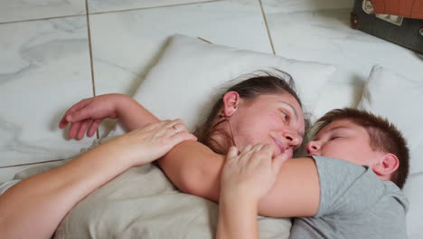 mother and son lying on white pillows resting together boy sleeping peacefully arms crossed around mother neck woman gazing upward wearing beige outfit marble tile floor visible cozy family moment