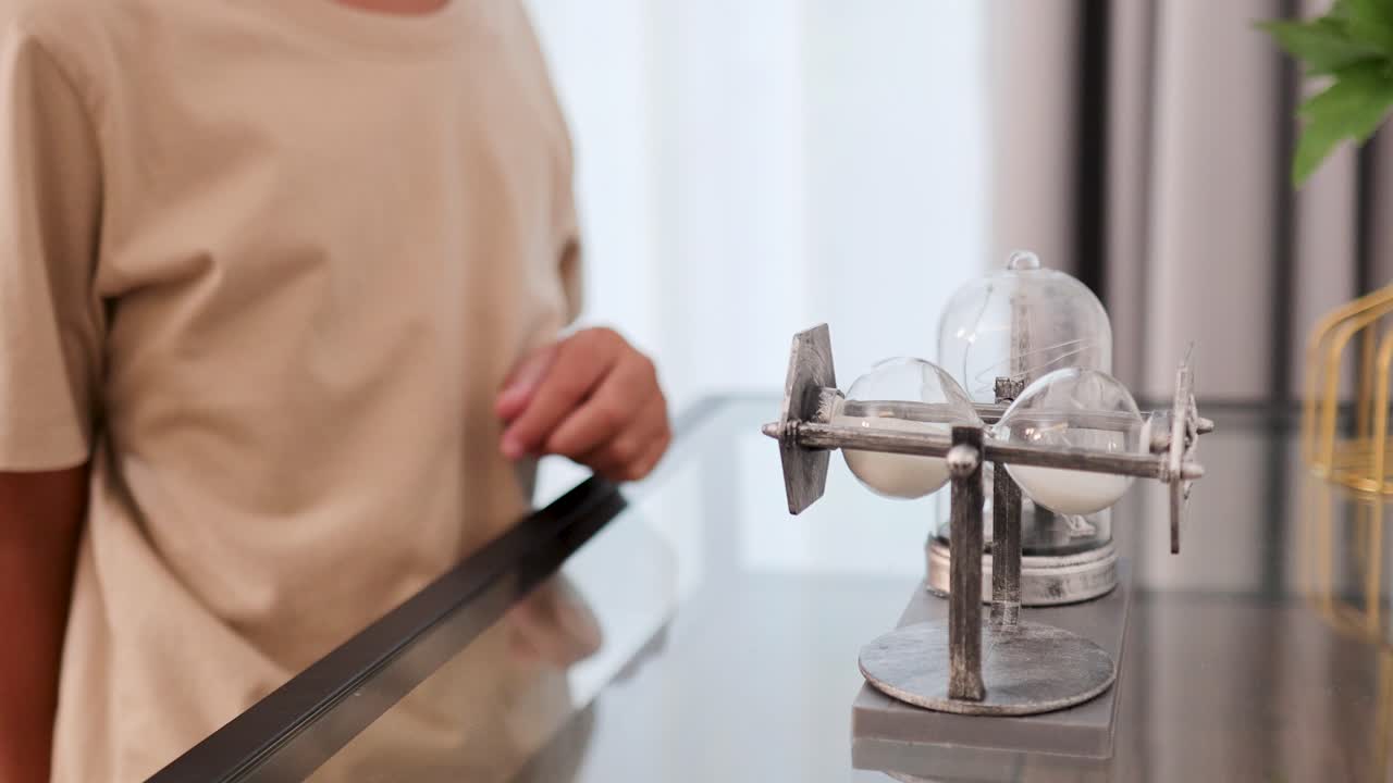 A child examines and rotates an hourglass timer on a table in a softly lit room, displaying curiosity and focus