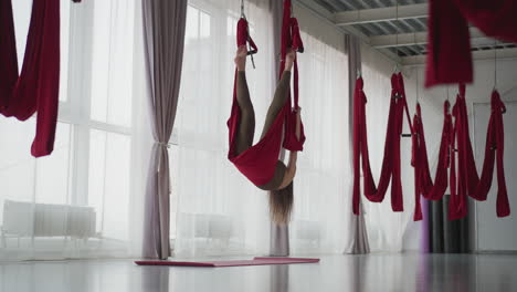 mindfulness practitioner gently swings in red aerial silk hammock by floor to ceiling window, legs suspended mid air, slight sway left right in bright modern studio conveying calm balance focus