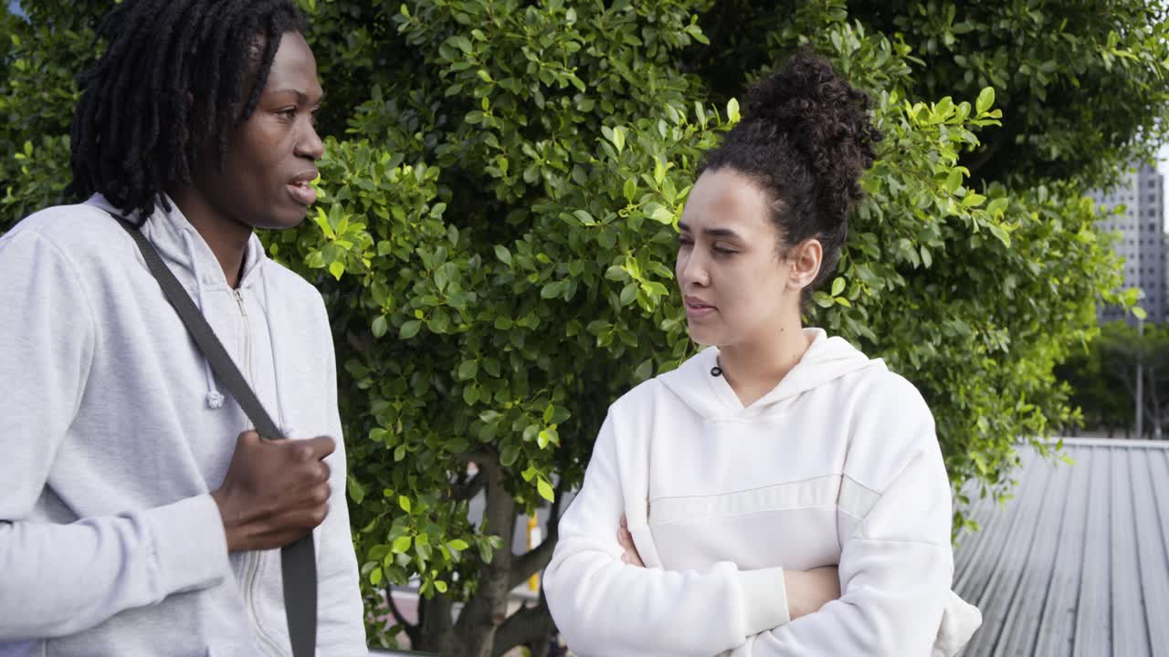 Uomo afroamericano che tiene la cinghia della borsa, iniziando una conversazione mentre una donna ascolta su un marciapiede accanto a un albero frondoso.