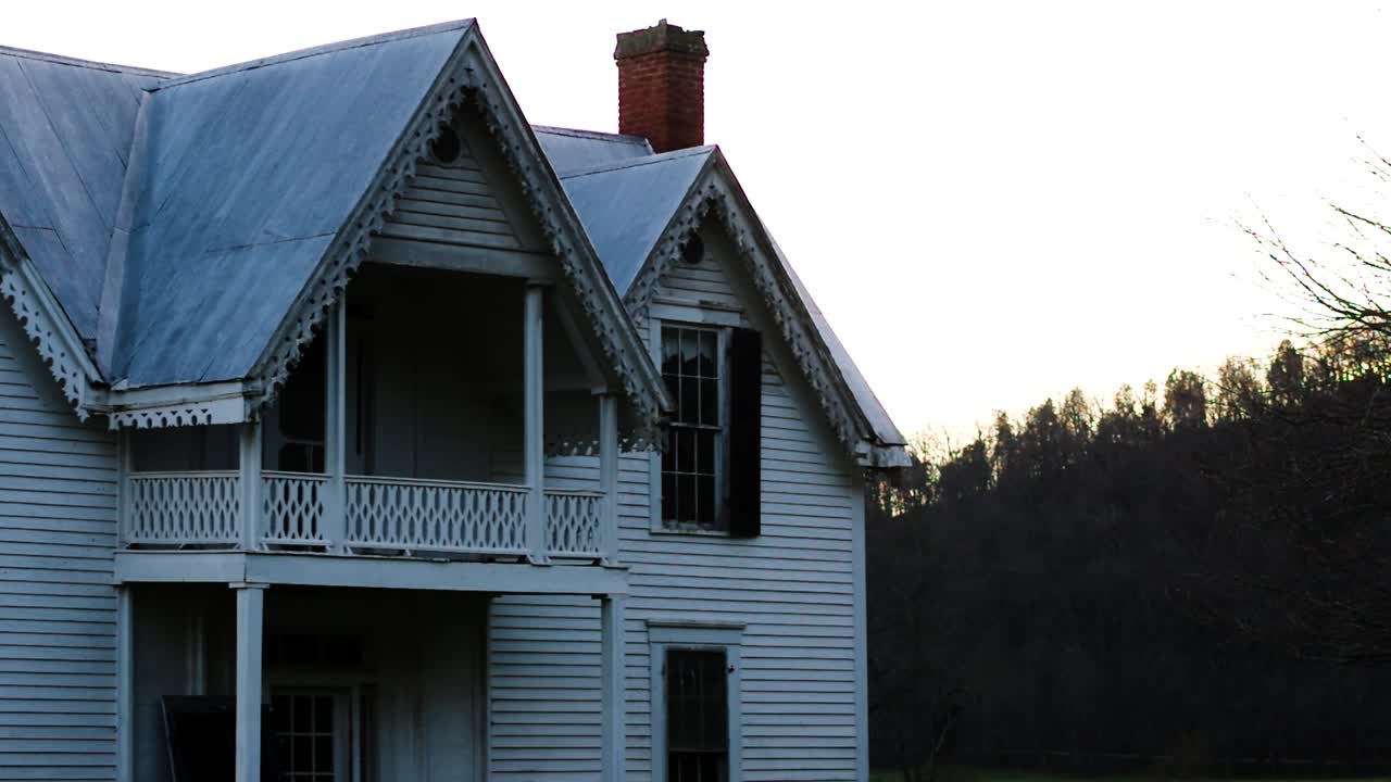 Abandoned older white home with decorative gables lined with mature trees with a spooky shadow backdrop