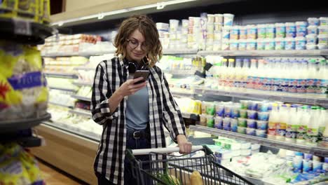 Concepto-De-Compras,-Tecnología,-Venta,-Consumismo-Y-Personas:-Mujer-Con-Teléfono-Inteligente-Y-Auriculares-En-El-Cuello-Caminando-En-El-Supermercado