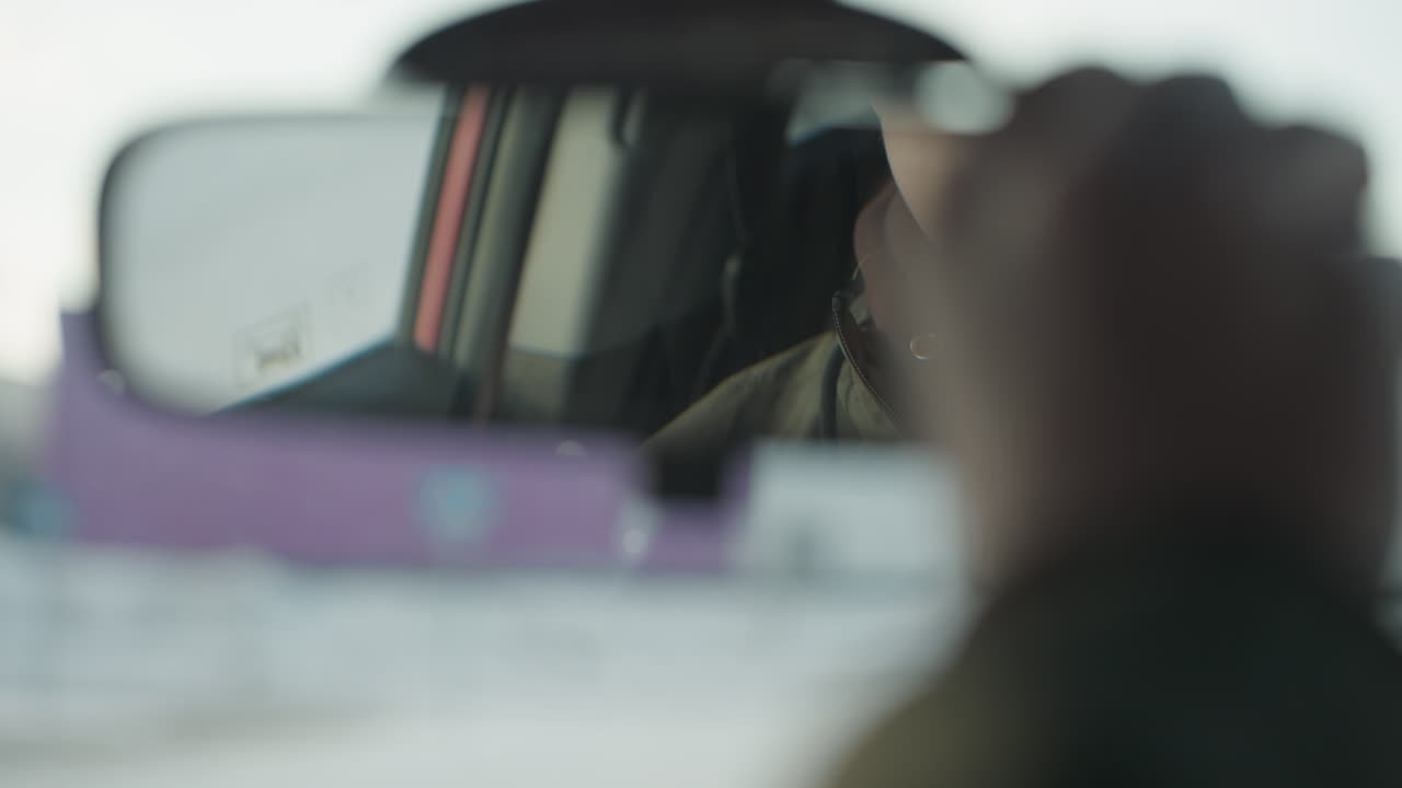 close up of rearview mirror inside car reflecting face of young boy adjusting mirror with one hand, wearing glasses, soft winter light visible in blur background through windshield