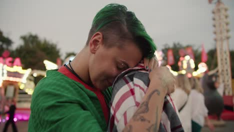 Close-up-shot-of-a-girl-with-short-green-hair-in-a-checkered-green-shirt-and-a-blonde-girl-with-a-short-haircut-in-a-checkered-pink-shirt-hugging-and-looking-at-each-other-during-their-date-in-the-amusement-park