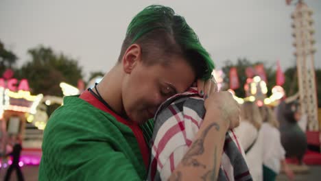 una foto en primer plano de una chica con cabello verde corto en una camisa verde a cuadros y una chica rubia con un corte de pelo corto en una camiña rosa a cuadros abrazándose y mirándose durante su cita en el parque de atracciones