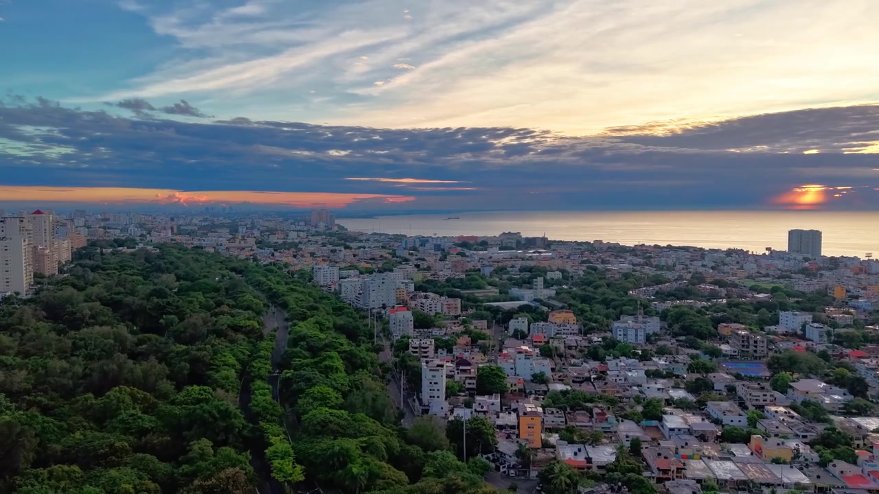 Hiperlapso aéreo de dron en ángulo alto volando hacia adelante sobre el denso dosel verde del Parque Mirador del Sur, mostrando nubes que se mueven rápidamente y luces de la ciudad desvaneciéndose durante un amanecer dramático en Santo Domingo