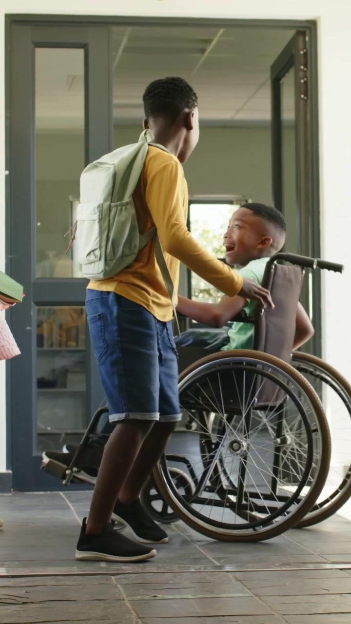 Video verticale: Che tiene la porta, aiutante in giallo che aiuta un amico in sedia a rotelle nell'atrio della scuola con uno zaino