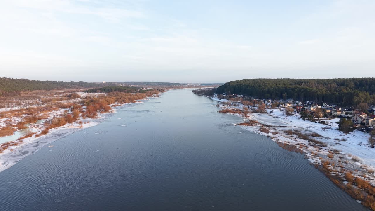 Dronevideo tallentaa upean ilmakuvan laajasta joesta, joka mutkittelee lumipeitteisen maiseman halki. Rauhallista näkymää kehystävät etäiset metsät kirkkaan taivaan alla