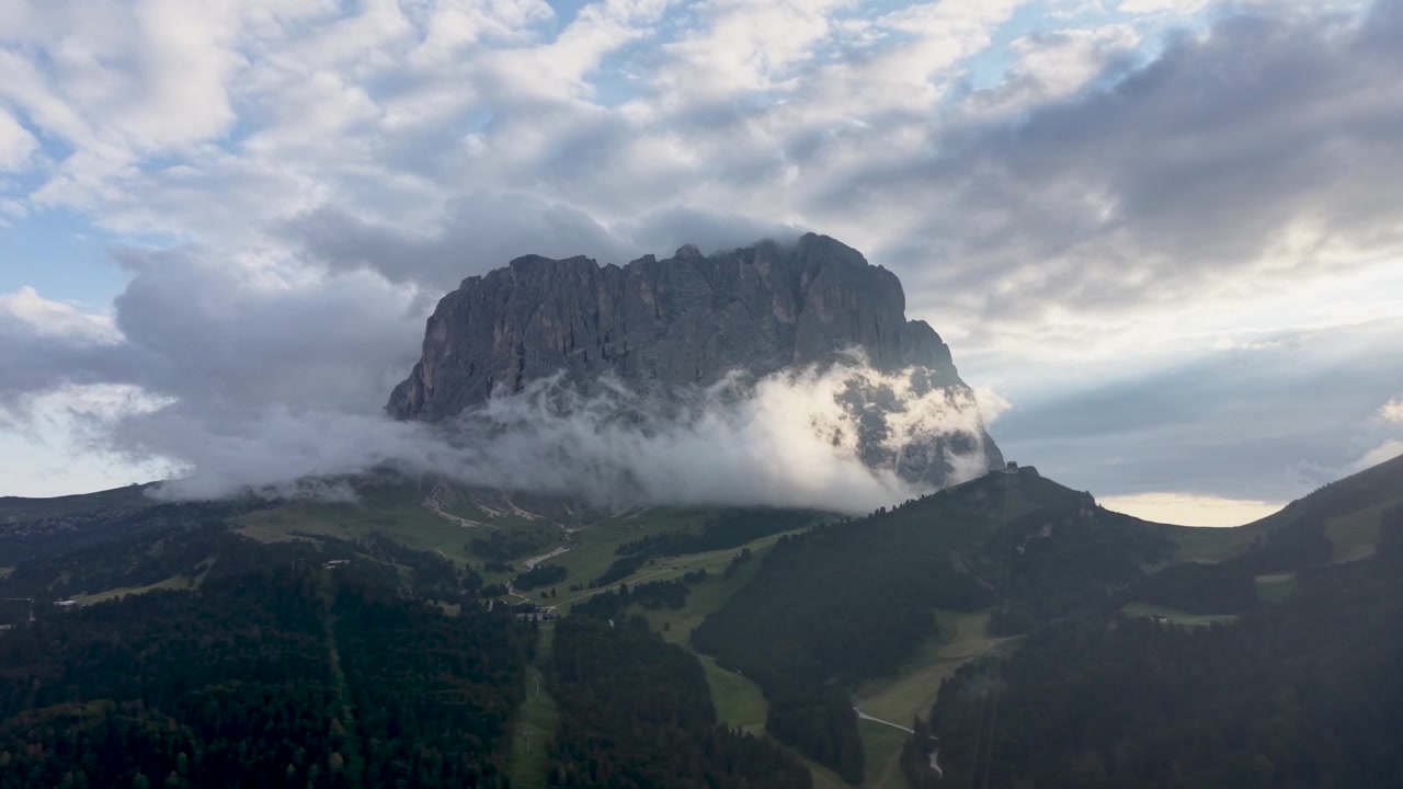 Vista aerea con drone del massiccio Gruppo del Sella circondato da nuvole e nebbia alla deriva, con verdi pendii alpini e terreno boscoso sotto una drammatica luce serale nelle Dolomiti.