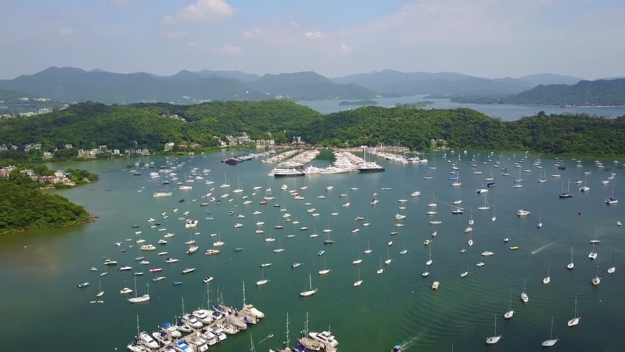 Drone video captures Discovery Bay marina in Hong Kong, showcasing hundreds of anchored boats against lush green hills and tranquil waters under a bright blue sky. Scenic beauty