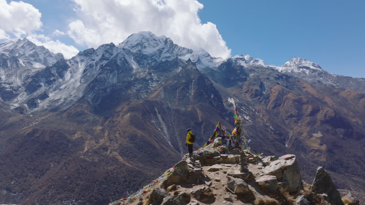 Drone shot reveals a male hiker standing on the edge of a high altitude summit in Nepal, panoramic view of the Himalayas Everest and Langtang mountain. Prayer flags flutter at vibrant, scenic setting.