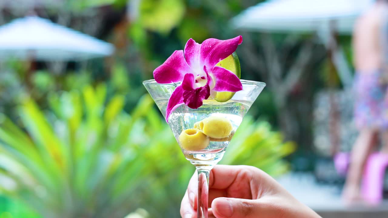 한 손 은 울창 한 열대 리조트 배경 에 꽃 을 장식 한 활기찬 마르티니 를 들고 있다