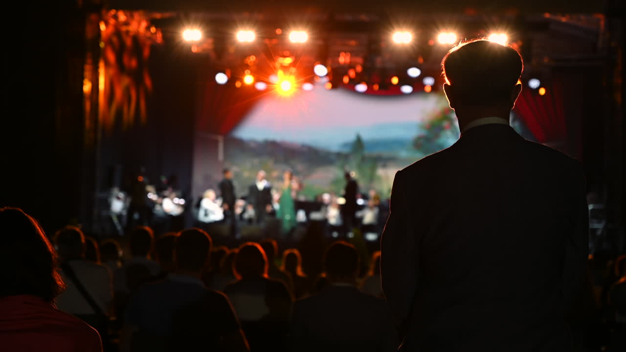 A lively performance takes place on stage with musicians and dancers, captivating an audience at an outdoor event in the evening. Colorful lights highlight the artistic display