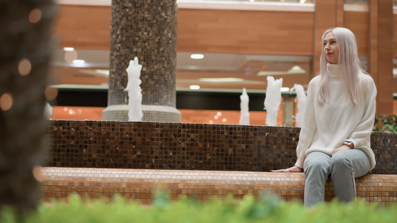 Sophisticated woman sits on mosaic bench by indoor fountain in shopping mall, calmly gazing around with gentle smile, ambient shoppers passing in background under warm lighting