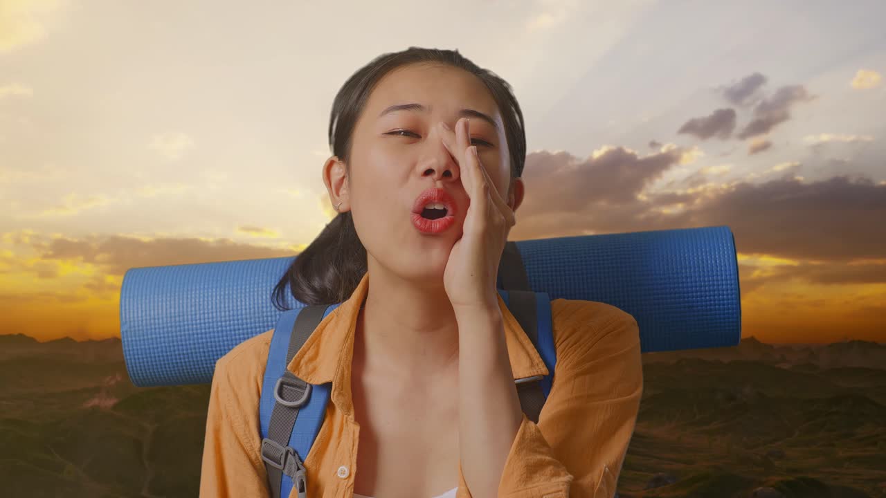 Close Up Of Asian Female Hiker With Mountaineering Backpack Screaming Holding Hands Near Her Mouth While Standing On The Top Of Mountain During Sunset Time