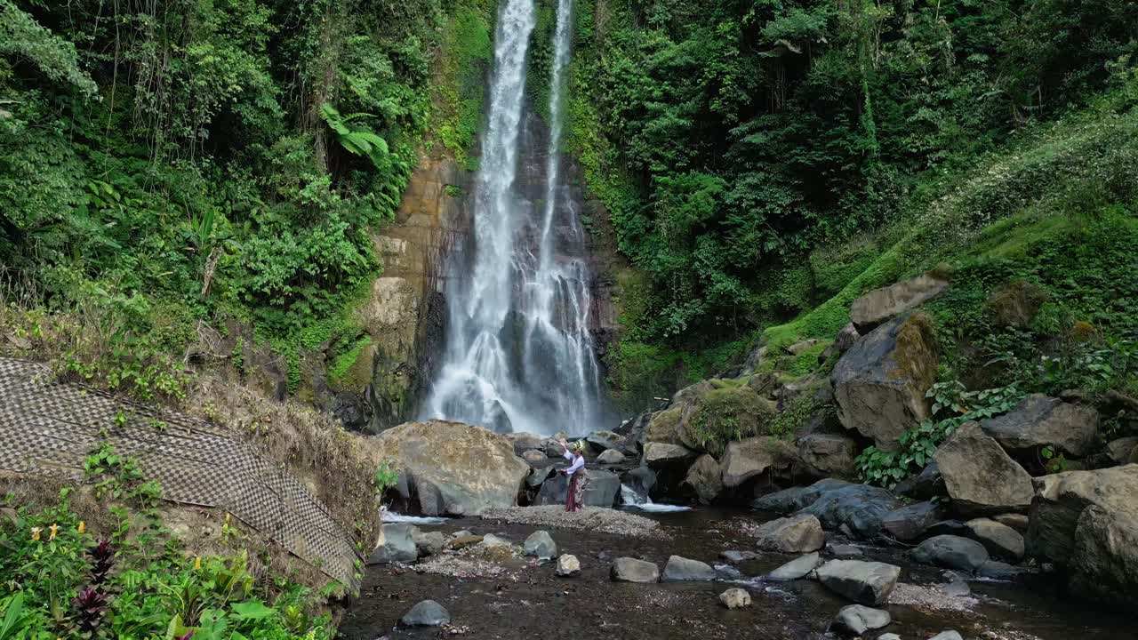 A Balinese female dancer performs gracefully in nature, wearing traditional attire and a leaf headpiece. Surrounded by greenery and shrubs, with Gitgit Waterfall visible in the background