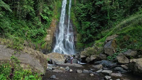 a balinese female dancer performs gracefully in nature, wearing traditional attire and a leaf headpiece. surrounded by greenery and shrubs, with gitgit waterfall visible in the background