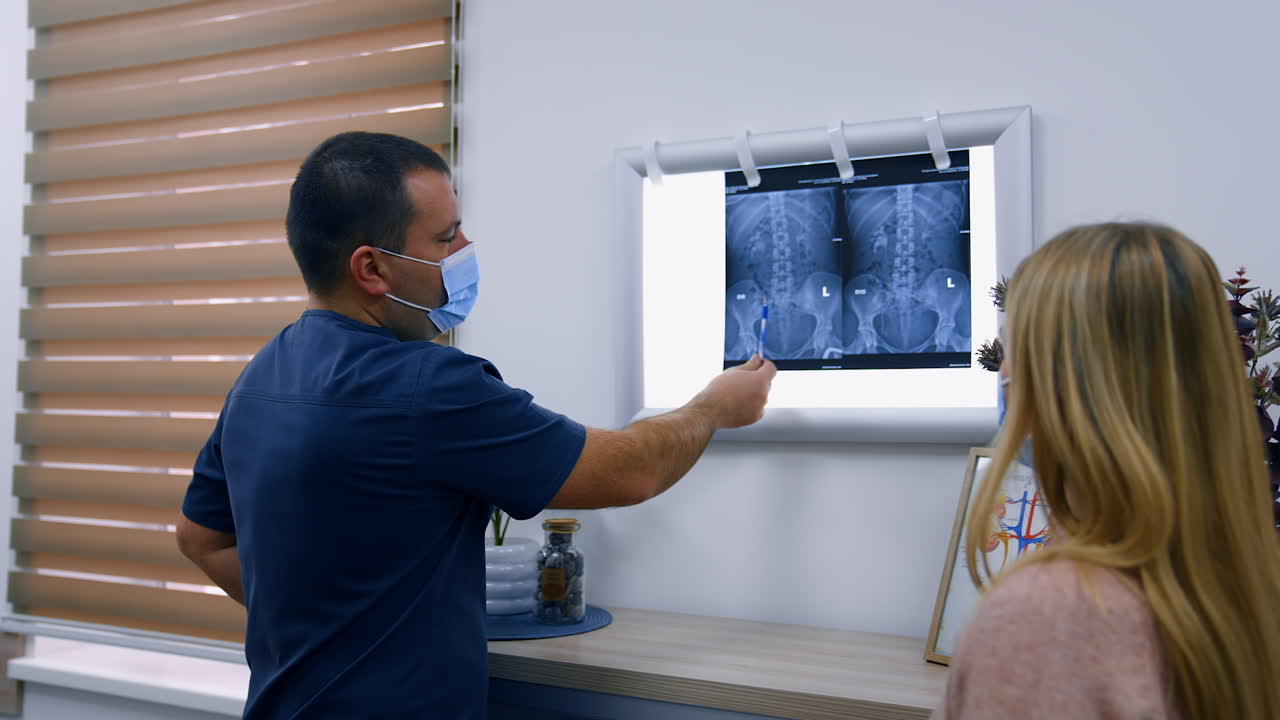 Mid-aged male medic wearing mask talks to patient pointing at x-ray scan. Health check up in modern hospital.