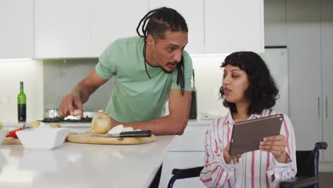 Happy-biracial-woman-in-wheelchair-using-tablet,-preparing-food-with-male-partner-in-kitchen