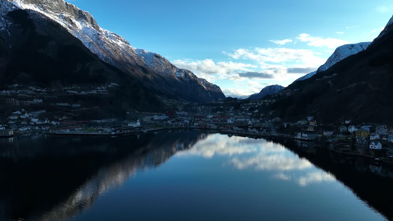 Snow capped peaks and glossy fjord in Odda Norway in clear winter evening. Valley in shadow, bright sky above
