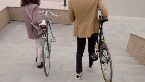 back view of american man and woman in formal clothes carrying their bikes down the stairs while going to work