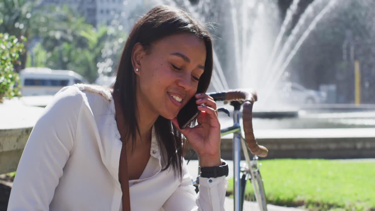 African american woman with bike talking on smartphone sitting in city park