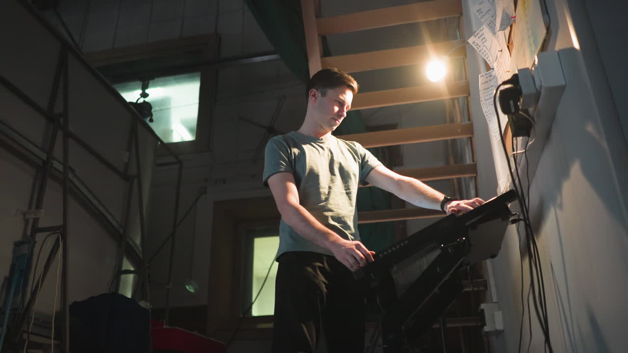Young technician stands in dim backstage room, staring at illuminated control panel under desk lamp near cork board filled with handwritten notes. Scene captures quiet focus in industrial workspace