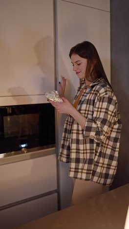 video vertical de una chica morena feliz en una camisa a cuadros comiendo por la noche recibiendo comida en papel de aluminio del refrigerador en la cocina en un apartamento moderno. una muchacha morena se levantó en medio de la noche y come comida en la cocina sacándola del refrigerador feliz y alegre