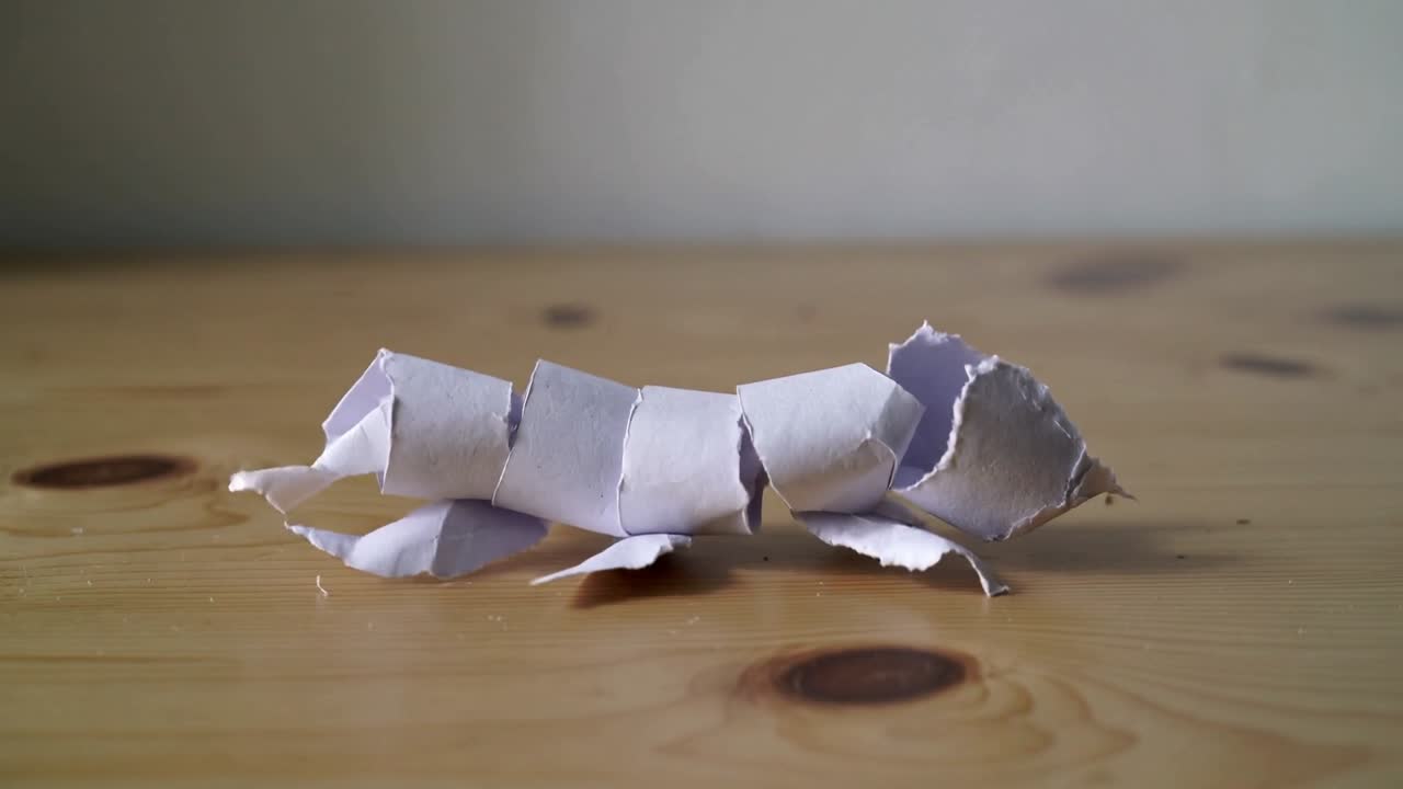 Frames iniciais: modelo de papel rasgado avançando sobre uma mesa de madeira, mostrando pedaços esfiapados.