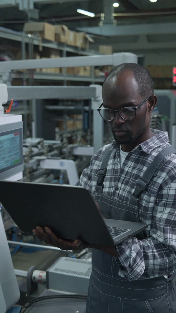 Black Factory Engineer Using Laptop to Control Automated Production Line