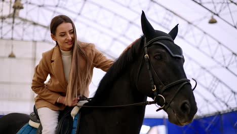 brunette woman sitting on a gorgeous black horse. lady leans forward to pet the animal. low angle view. manege at backdrop.