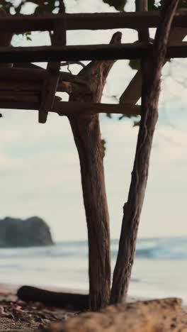 weathered wooden structure made of tree branches stands on a beach, overlooking calm ocean waves and a distant rocky coastline