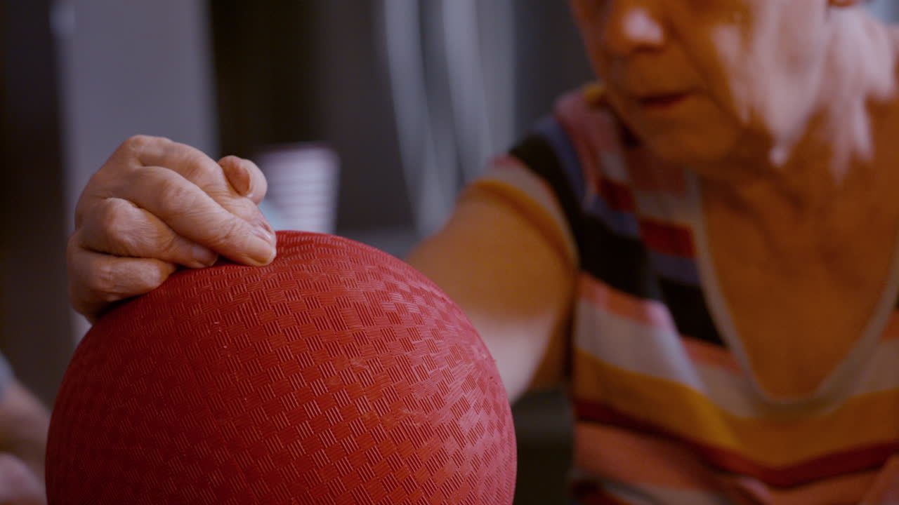 Male caregiver gently assists the hemiplegic right hand ball therapy of an elderly female stroke survivor, seated across from him on glass top kitchen table at home. Side to side motion of hand 24fps.