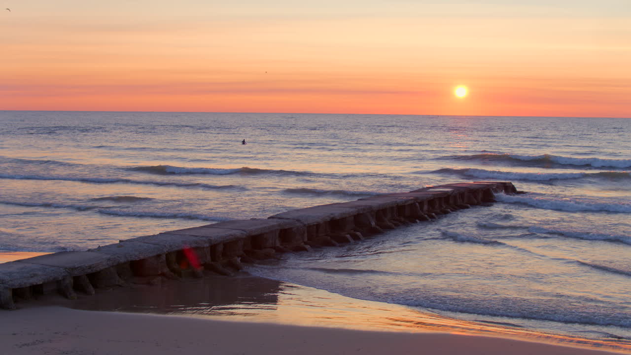 Drone aerial over Lake Michigan at sunrise with a surfer floating in gentle waves near a pier, pulling back to reveal sandy beach and glowing horizon