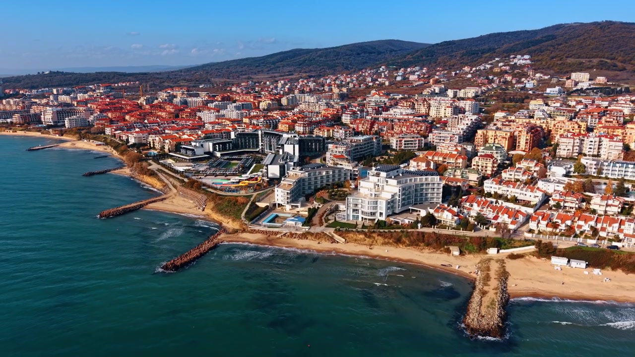 Veduta aerea panoramica di una località balneare con hotel lungo la costa. Ampia ripresa con drone di edifici moderni e residenziali sul fianco della collina, con spiagge sabbiose e frangiflutti.