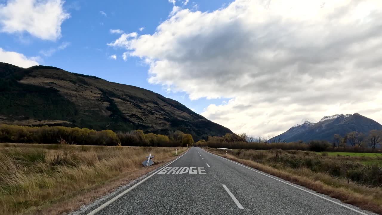 Forward-moving car view along rural road, mountains, autumn trees, dynamic clouds, natural daylight