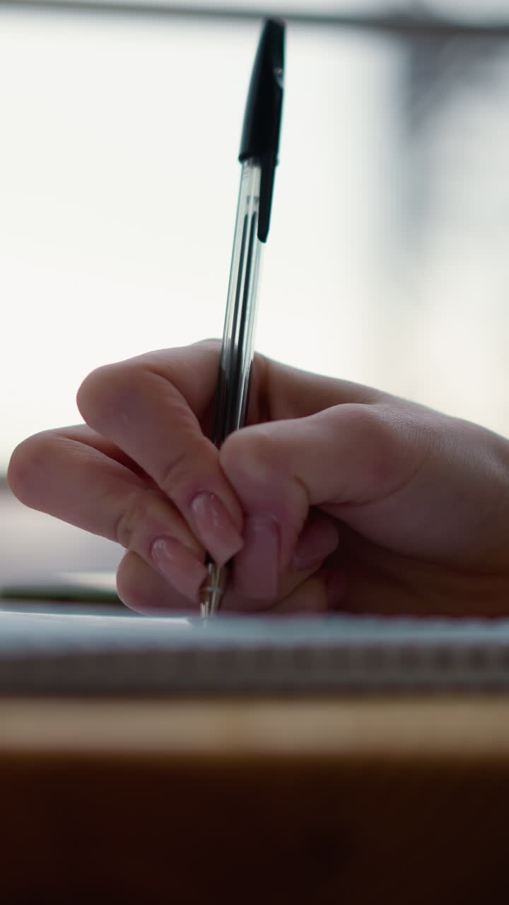Close-up of hand writing in notebook on wooden table with beverage cup and blurred background showcasing structure, creating a cozy and focused work or study environment