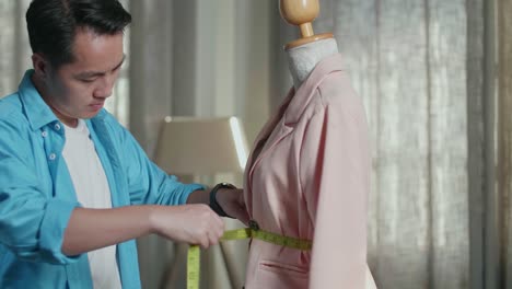 primer plano de un diseñador masculino asiático con una máquina de coser midiendo una chaqueta en un maniquí mientras diseña ropa en el estudio