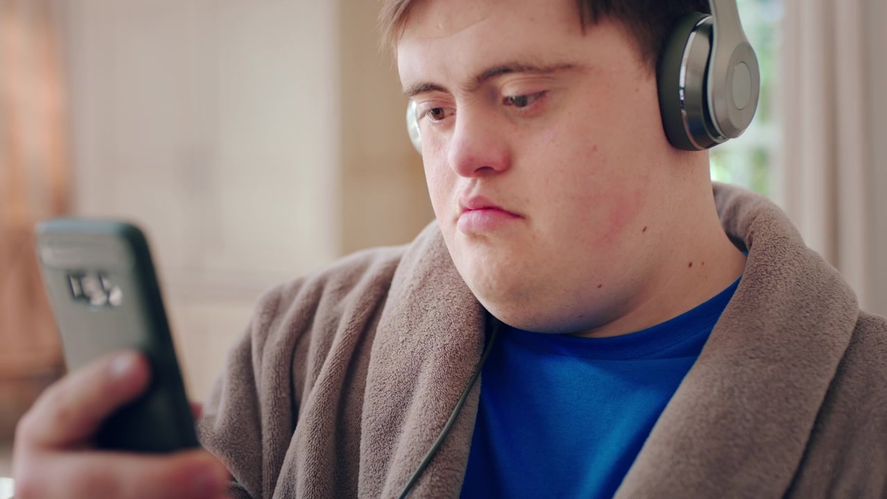 Ung mand med Downs syndrom bruger smartphone og hovedtelefoner derhjemme