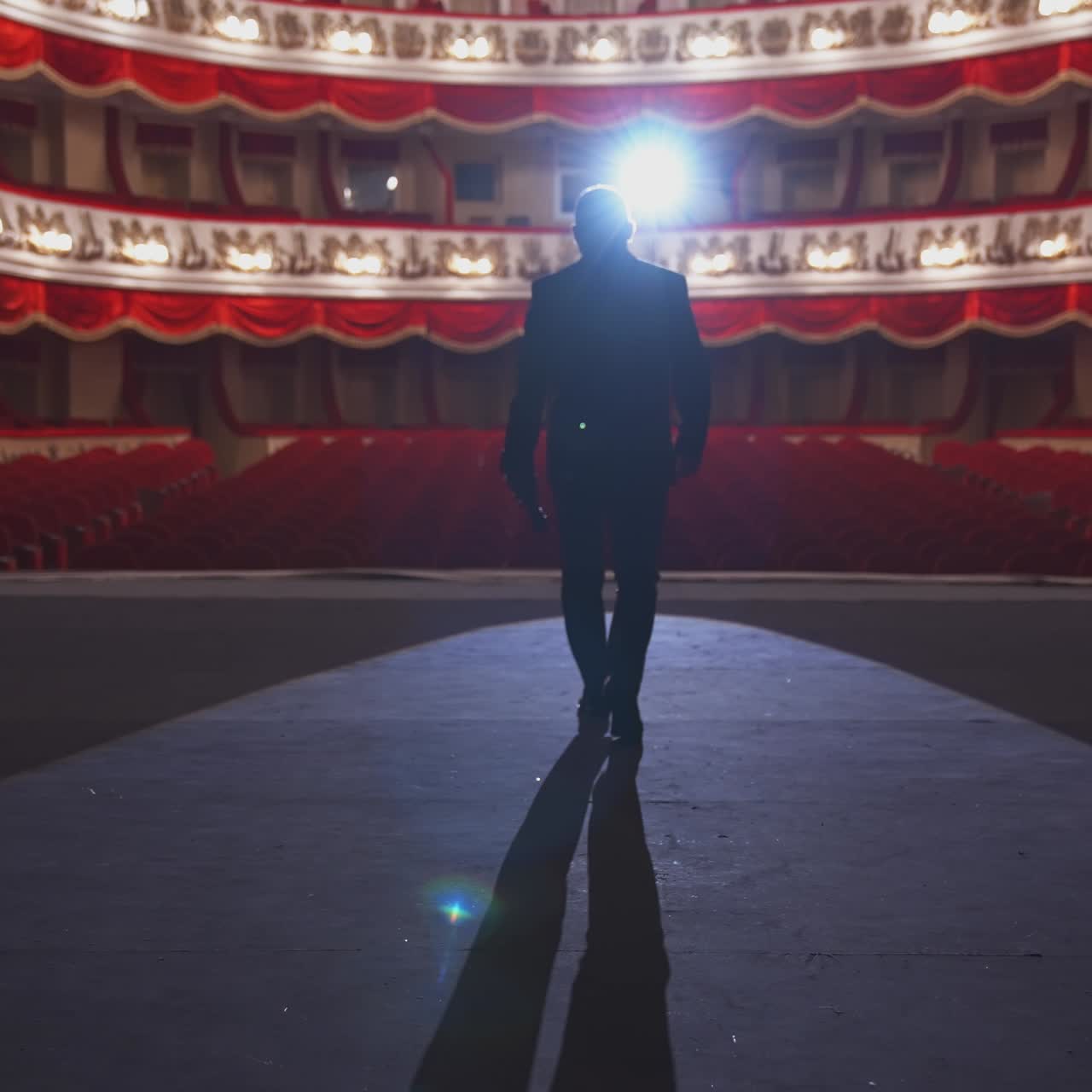 Man with microphone on theatre stage. Male actor in elegant suit walks on scene and starts speech in front of the empty auditorium. Back view.