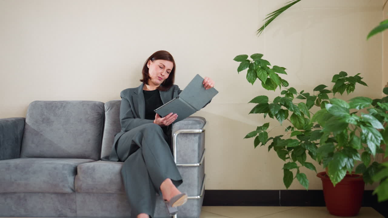 Confident businesswoman in gray suit sitting cross-legged on modern sofa using tablet in bright indoor office lounge with green potted plants, focused on screen in calm, professional environment