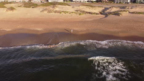 Man-walking-alone-on-beach-at-sunrise-with-city-buildings-in-background,-Punta-del-Este-in-Uruguay